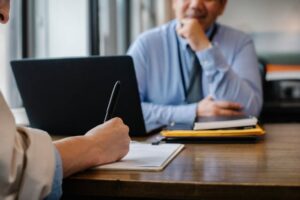 Business professional taking notes during a meeting with a colleague in a modern office setting, with documents and a laptop on the table.